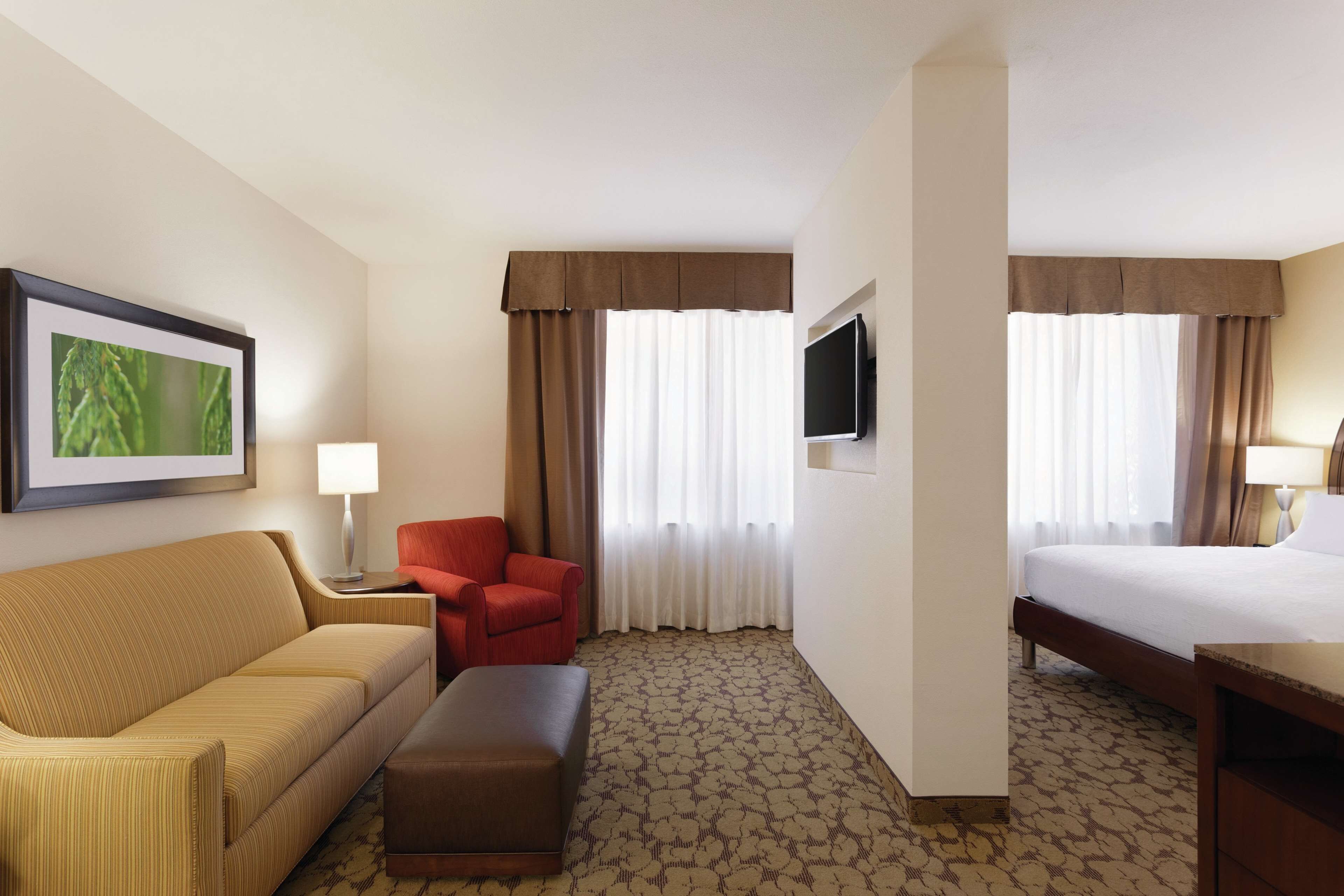 Habitación de hotel con cama, sofá, sillón rojo, una mesita de noche con lámpara y televisión en la pared.