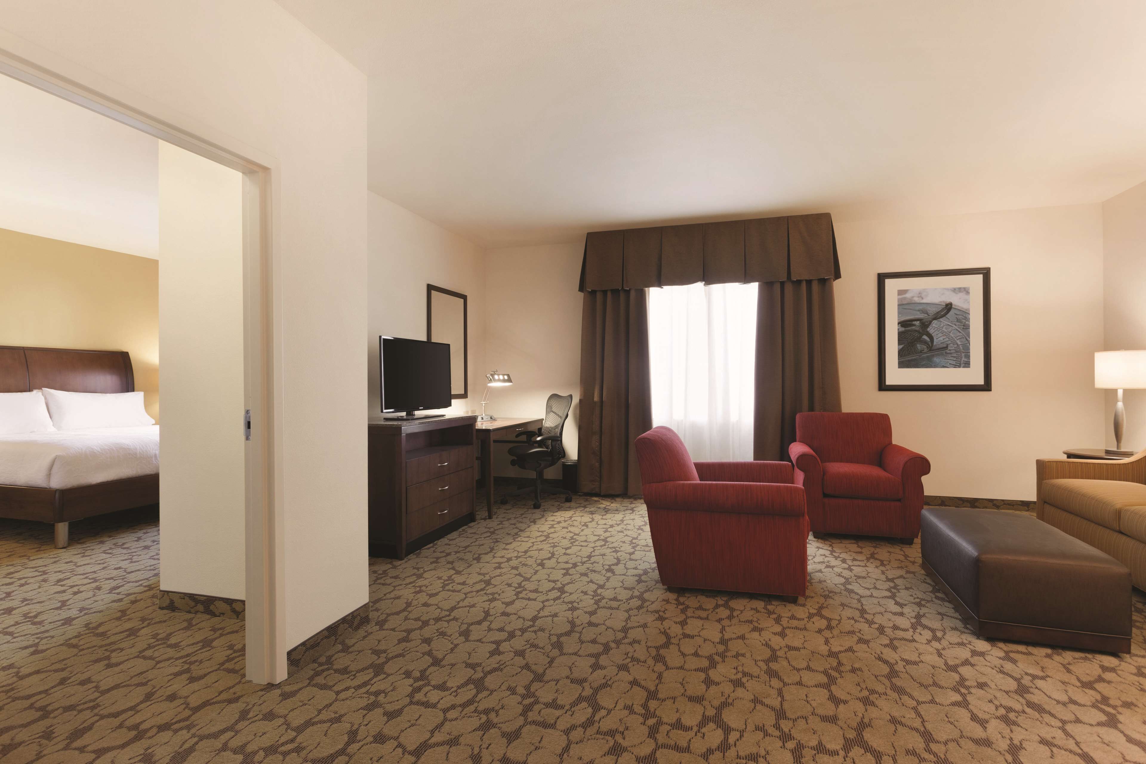 Ein Hotelzimmer mit einem Bett, einem Schreibtisch, einem Fernseher, einem roten Sessel, einem braunen Hocker und einem Fenster mit Vorhängen.