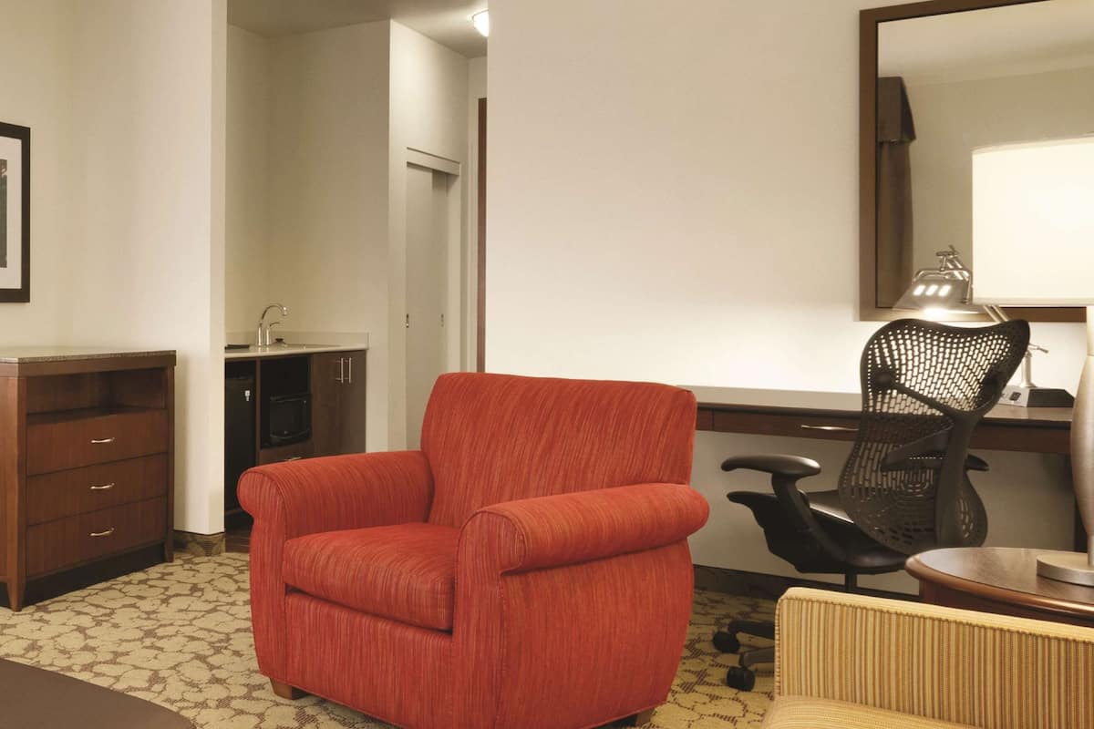 A hotel room with a red armchair, a wooden cabinet, a desk with a chair, a lamp, and a mirror.