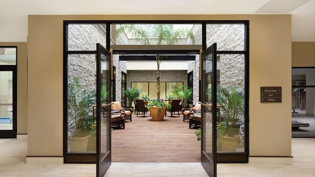 An indoor area with a wooden floor, glass doors leading to a space with chairs and potted plants. - Hilton Garden Inn Rochester Downtown, NY (Rochester)