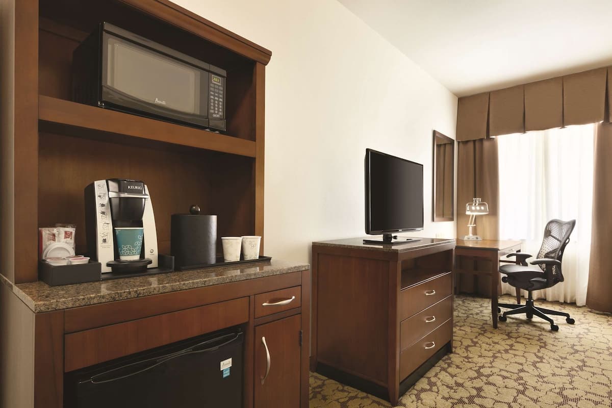 A hotel room with a kitchenette, including a microwave, coffee maker, and refrigerator. There is a TV on a wooden stand and a desk with a chair.