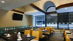A hotel dining area with tables and chairs, a television on the wall, and large windows with a view of the street.