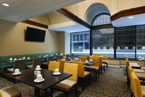 A hotel dining area with tables and chairs, a television on the wall, and large windows with a view of the street.