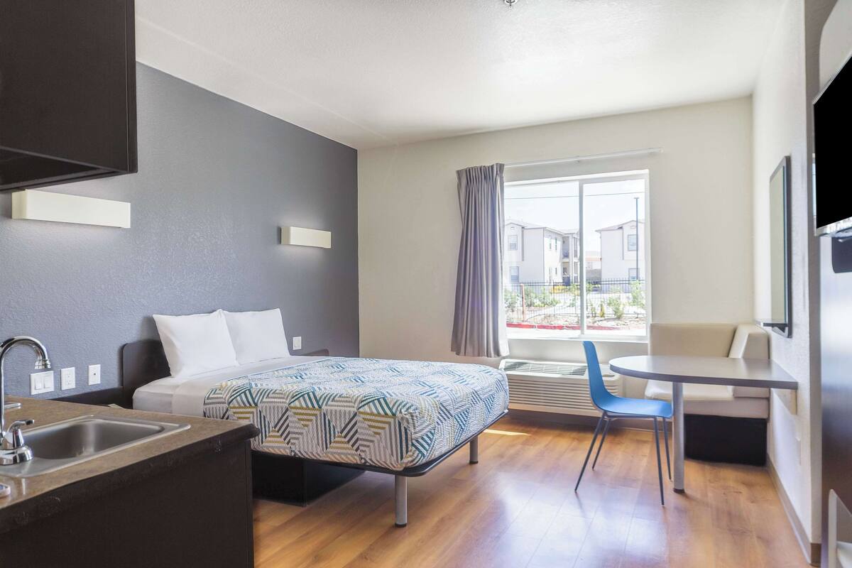 A modern hotel room with a bed, a desk, a chair, a sink, and a large window with curtains.