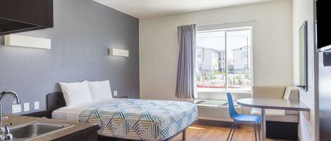 A modern hotel room with a bed, a desk, a chair, a sink, and a large window with curtains.