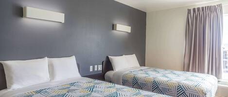 A hotel room with two beds, each with a patterned bedspread, a grey wall, and a window with curtains.