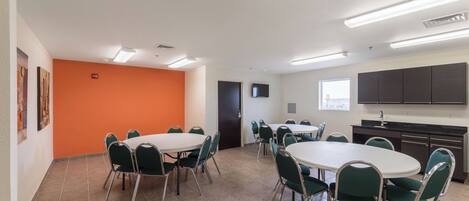 A conference room with a white table and green chairs, an orange accent wall, a TV, and a kitchenette with dark cabinets.
