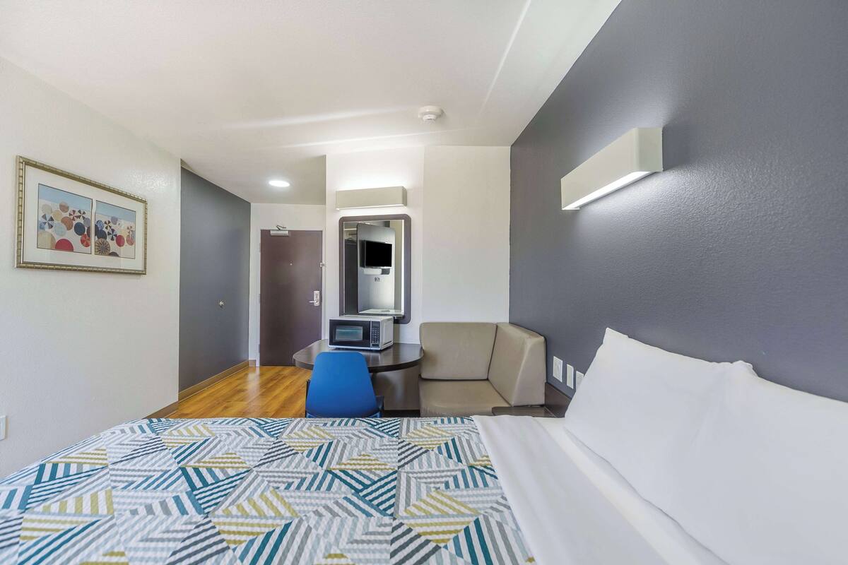 A hotel room with a bed, a desk with a chair, a microwave, a TV, and a door leading to another room.