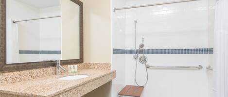 A bathroom with a granite countertop sink, a mirror, a shower with a grab bar, and a towel rack.
