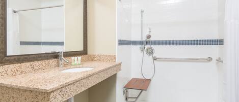 A bathroom with a granite countertop sink, a mirror, a shower with a grab bar, and a tiled floor.