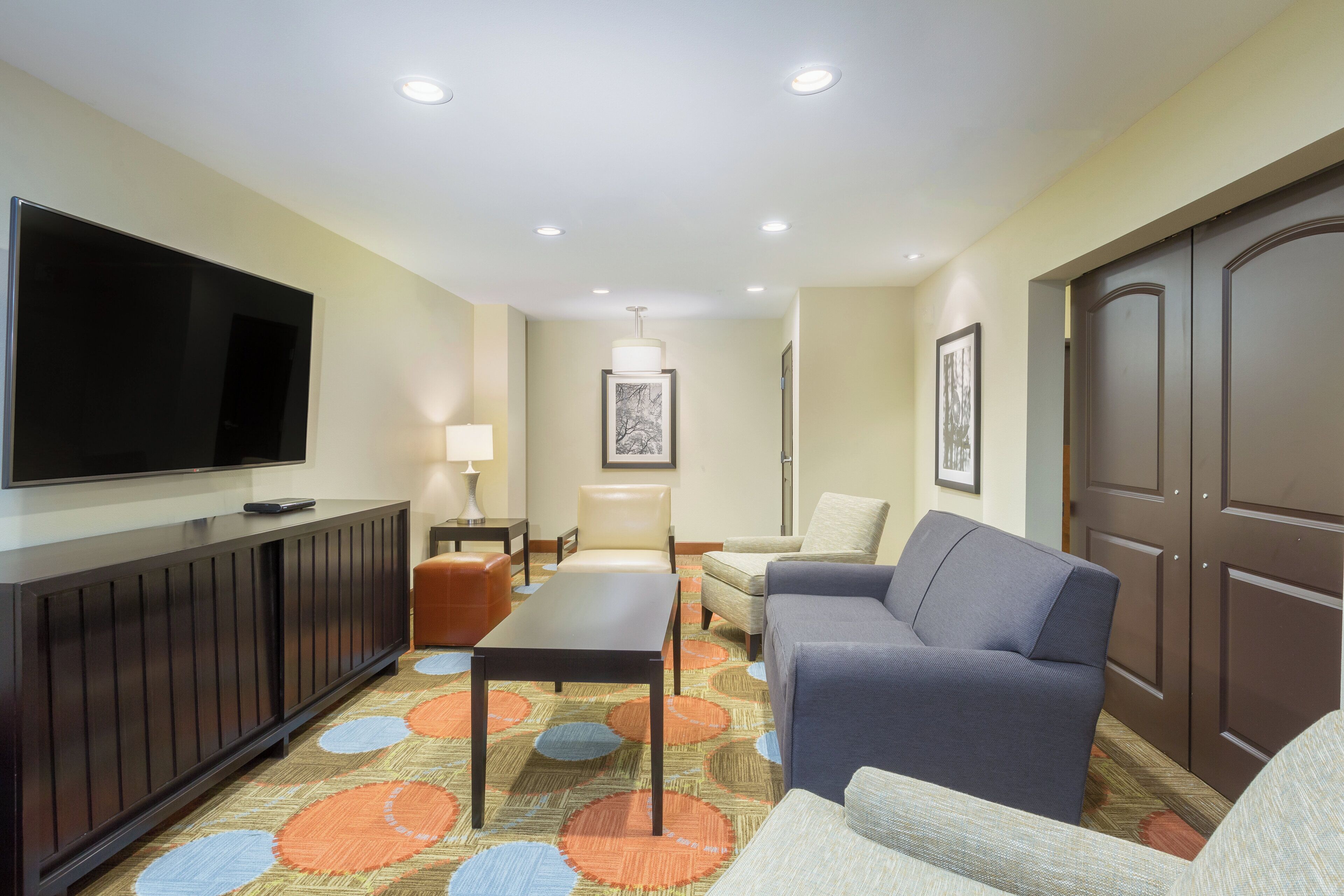 A hotel room with a flat-screen TV, a sofa, a chair, a small table, and a patterned carpet.