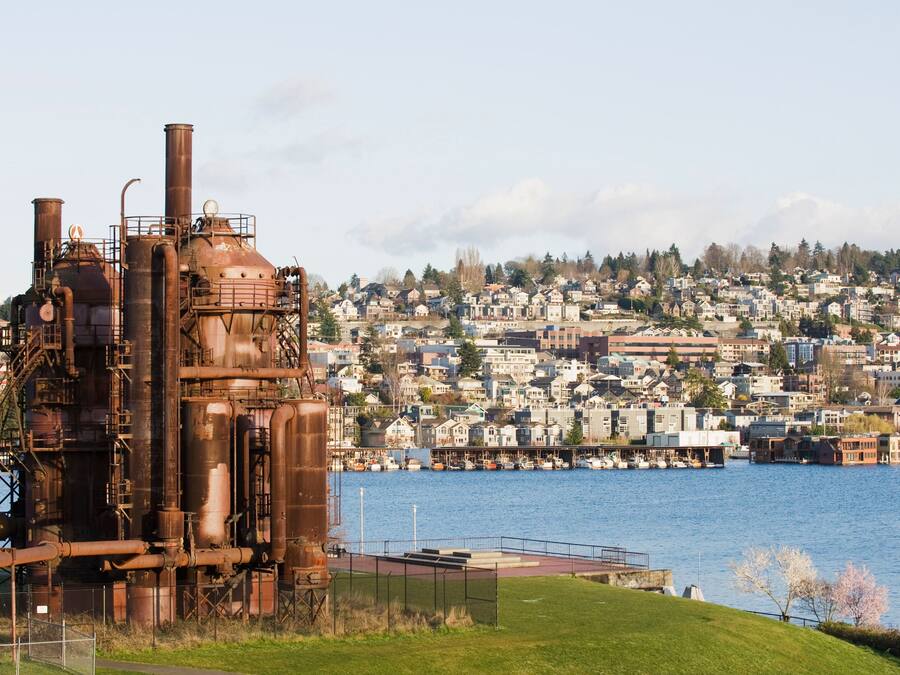 An industrial structure with multiple chimneys and pipes, a waterfront with boats, and a densely built residential area in the background.