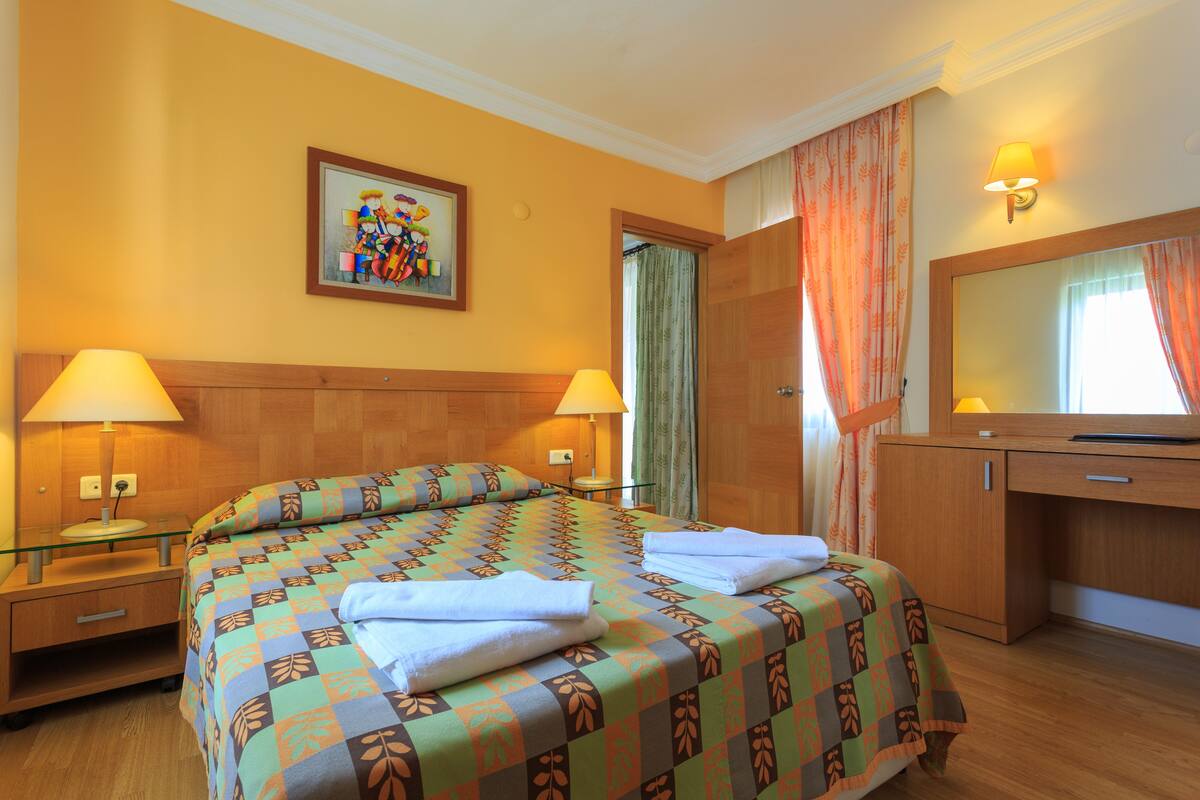 Une chambre d’hôtel avec un lit, deux lampes de chevet, une table de nuit en bois, un bureau avec un miroir et un tableau au mur.