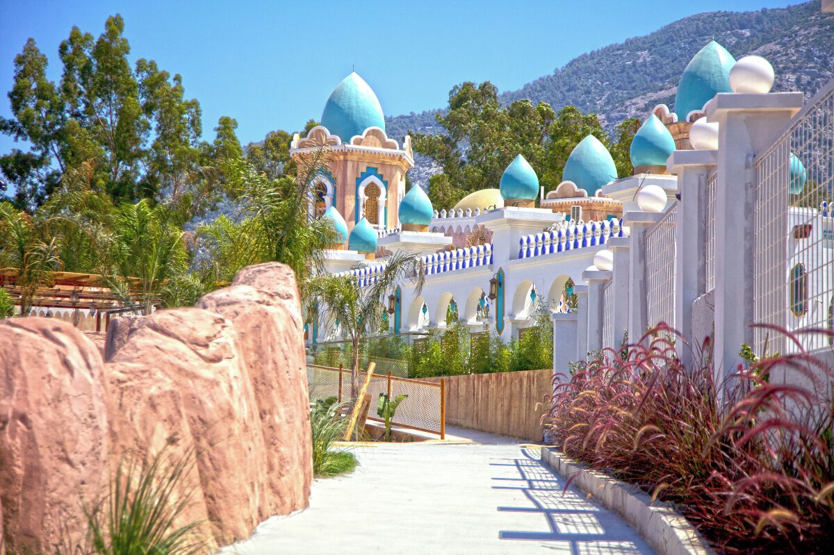 A resort with a central building featuring domed roofs, surrounded by a white fence and palm trees.