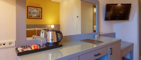 Camera d'albergo con una scrivania in vetro, una TV a schermo piatto montata a parete, uno specchio grande sopra la scrivania e un vassoio con bollitore e tazze da caffè.
