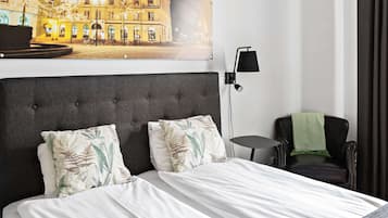 Ett hotellrum med en stor säng, två kuddar, ett nattduksbord, en lampa, en stol och ett konstverk i form av en bild av en byggnad som hänger på väggen.