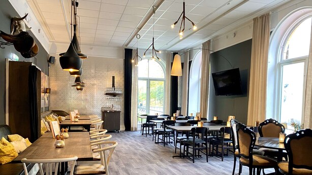 Un restaurant avec des tables et des chaises en bois, des lampes suspendues et une grande fenêtre ornée de rideaux.