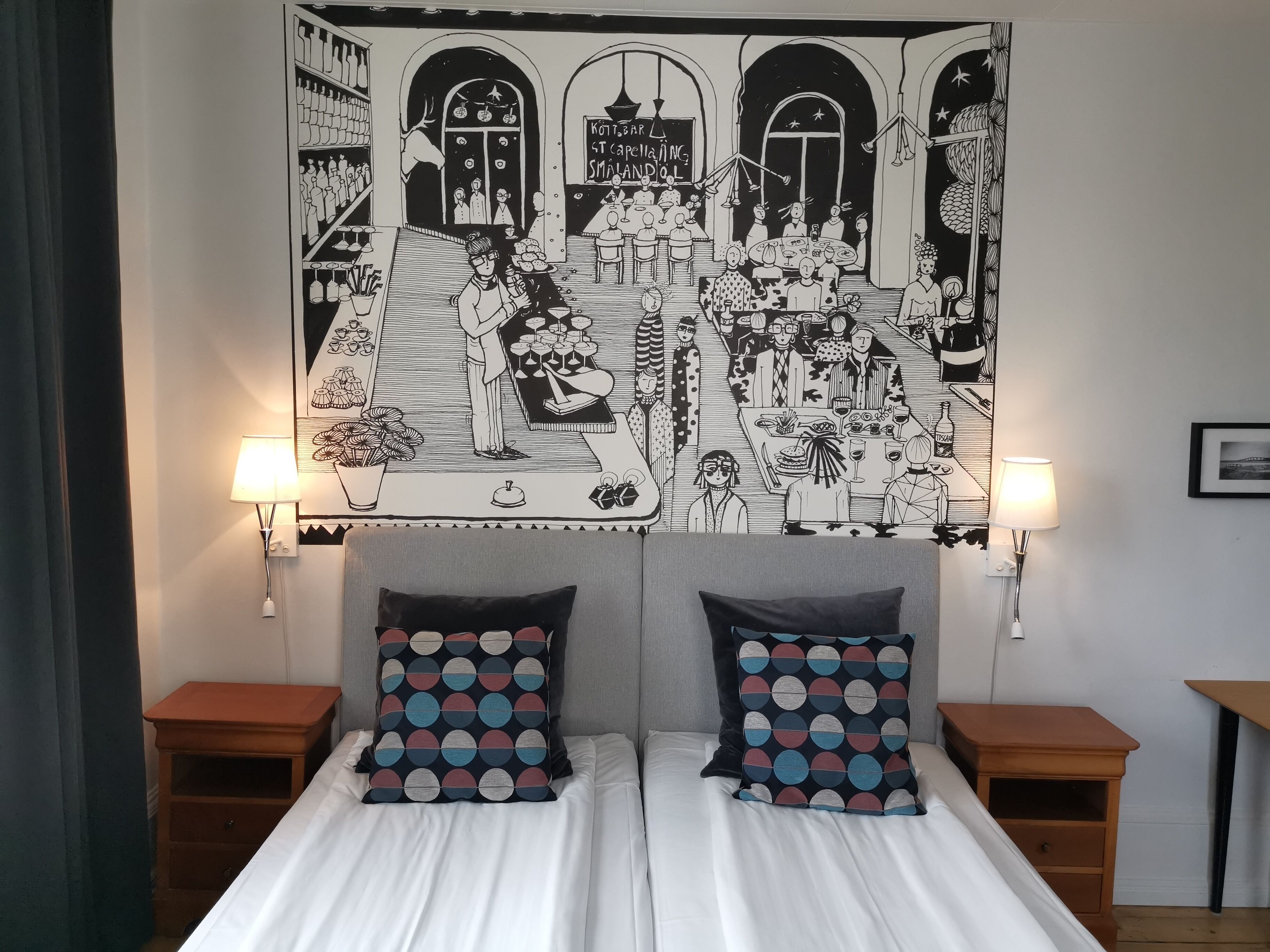 A bedroom with a large black and white tapestry depicting a social scene, two bedside tables with lamps, and a bed with patterned pillows.