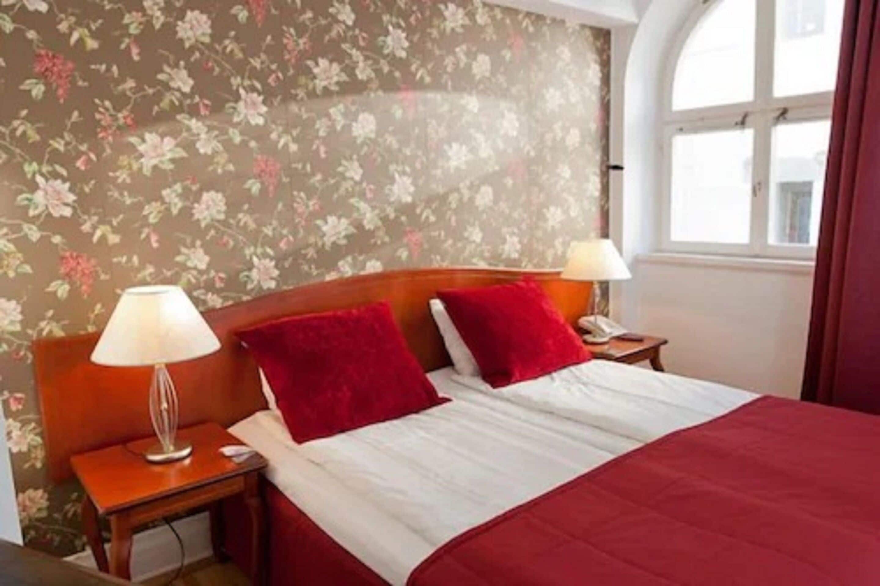 Una habitación de hotel con papel tapiz floral, un cabecero de madera, una cama con ropa de cama roja y blanca, dos lámparas de noche y una ventana con cortinas.