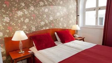 Ein Hotelzimmer mit floralem Wandpapier, einem hölzernen Kopfteil, einem Bett mit rot-weißer Bettwäsche, zwei Nachttischlampen und einem Fenster mit Vorhängen.