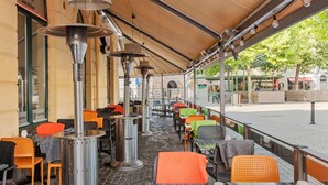Un café en plein air avec des tables et des chaises, un espace de repos abrité et, en arrière-plan, une vue sur la rue.