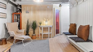 Une pièce chaleureuse avec un mur en bois, un canapé blanc agrémenté de coussins, un fauteuil en osier, une petite table avec une lampe et une plante en pot.