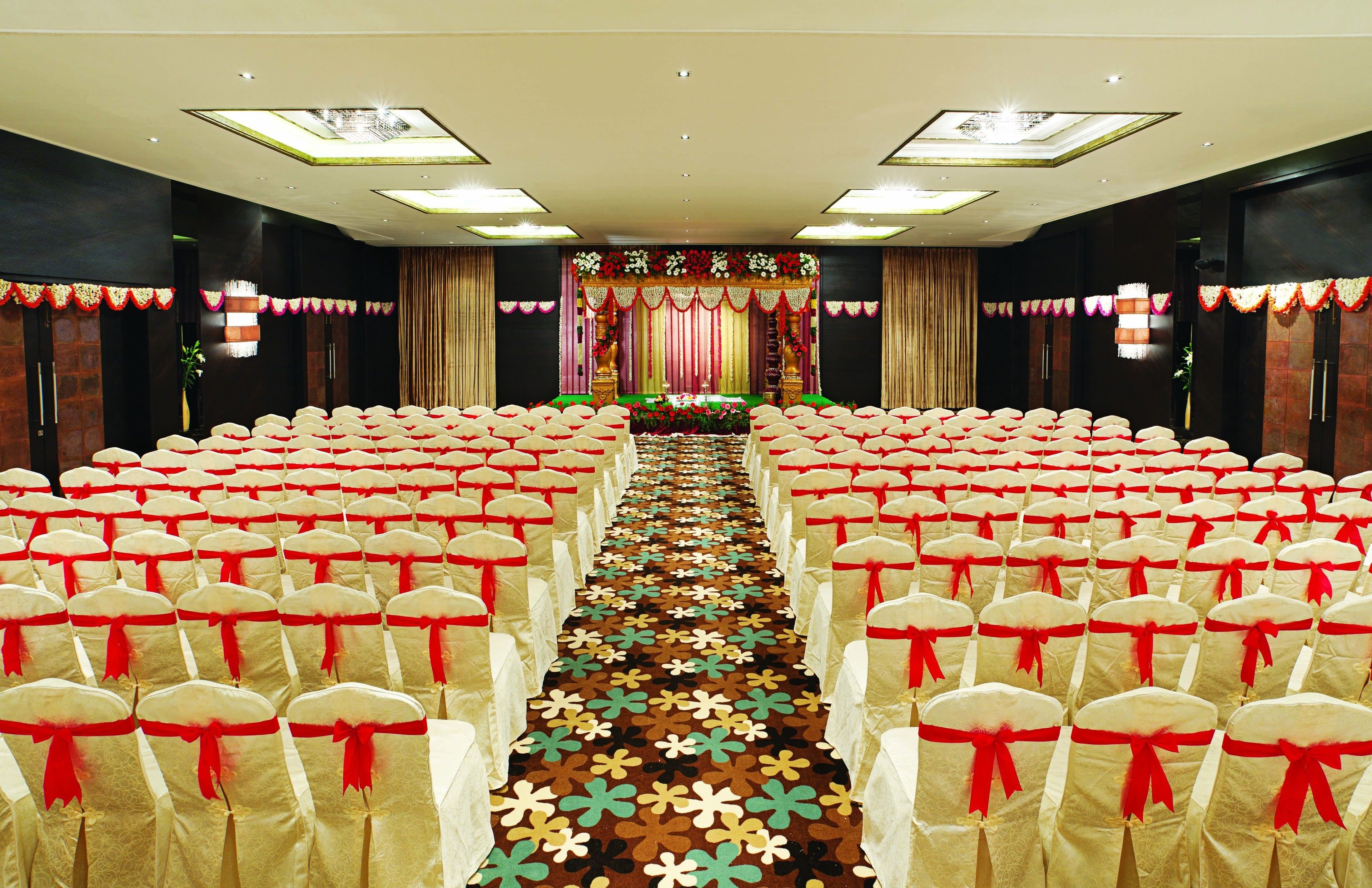 A conference room with rows of chairs, a stage with floral decorations, and a carpet with a floral pattern.