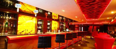 Zona de bar con una decoración en tonos rojo y amarillo, varios taburetes y una variedad de botellas y vasos en estanterías.