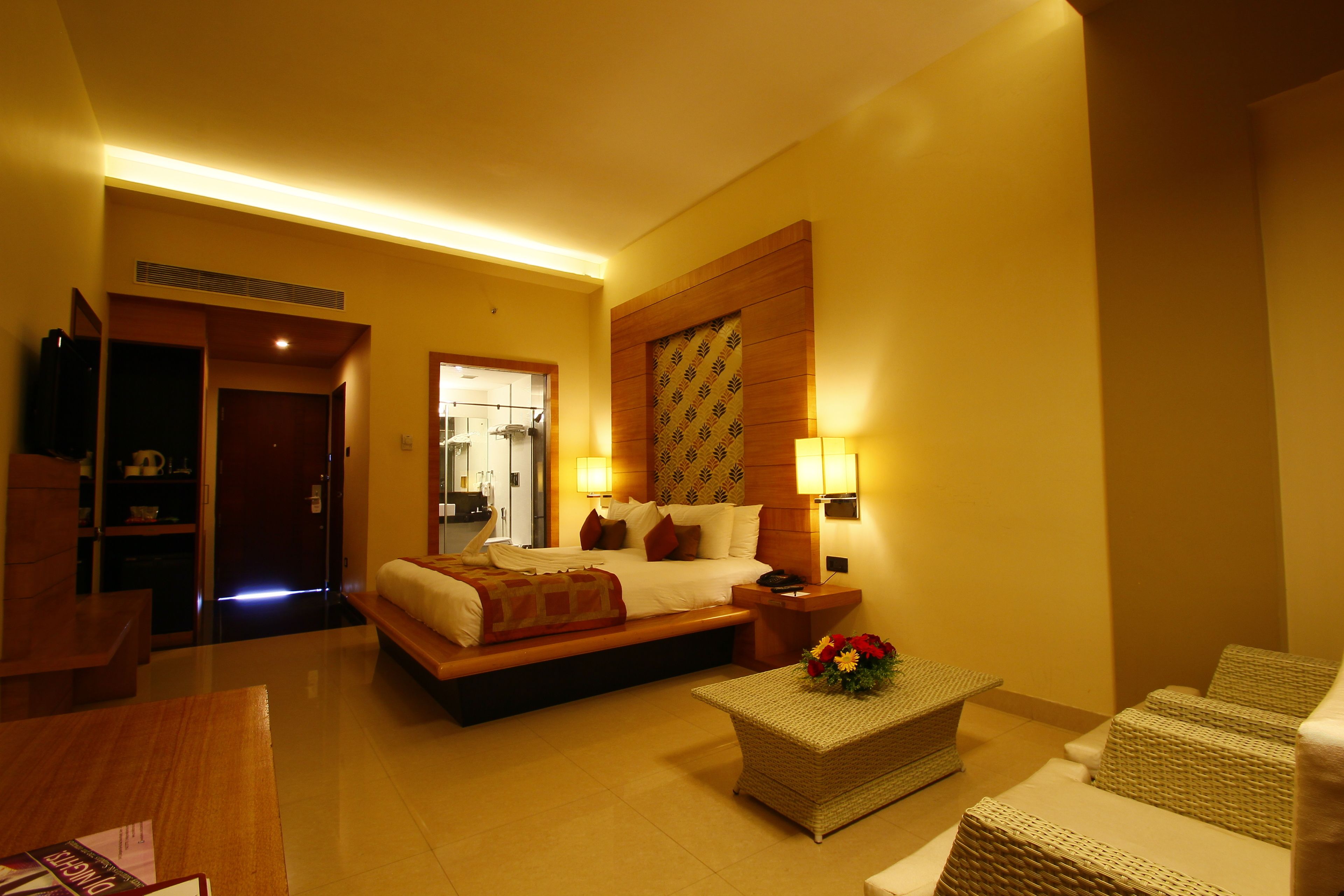 A hotel room with a bed, bedside tables, a small sofa, a coffee table with flowers, and a bathroom visible through an open door.