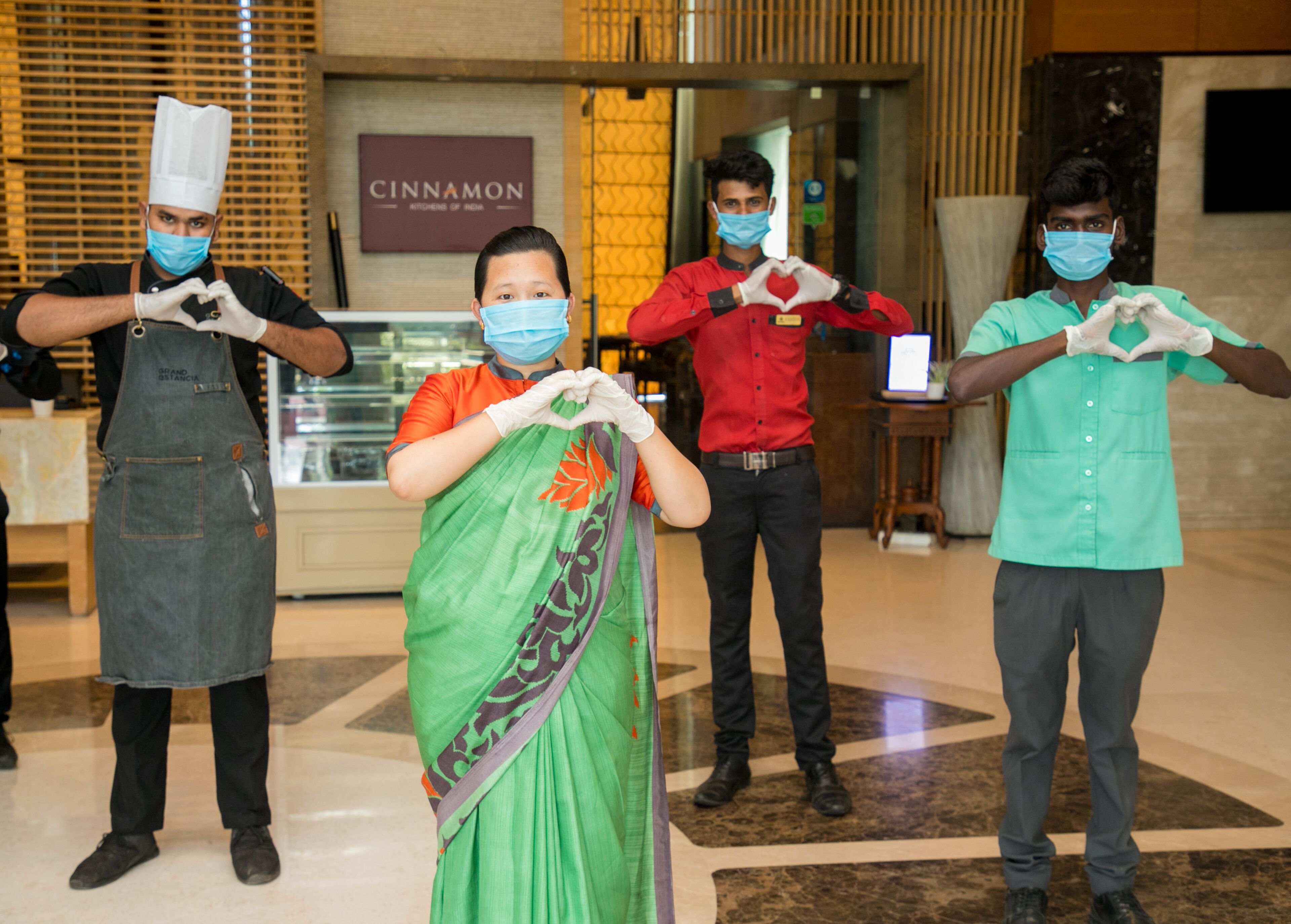 Four individuals in a lobby, one in a chef's hat and apron, another in a green sari, and two in casual attire, all making heart shapes with their hands.