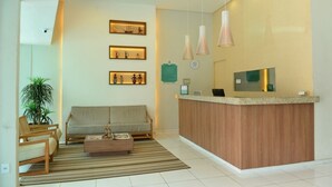 A reception area with a wooden reception desk, a sofa, a coffee table, and a potted plant. - Alpha Praia Hotel (Fortaleza)