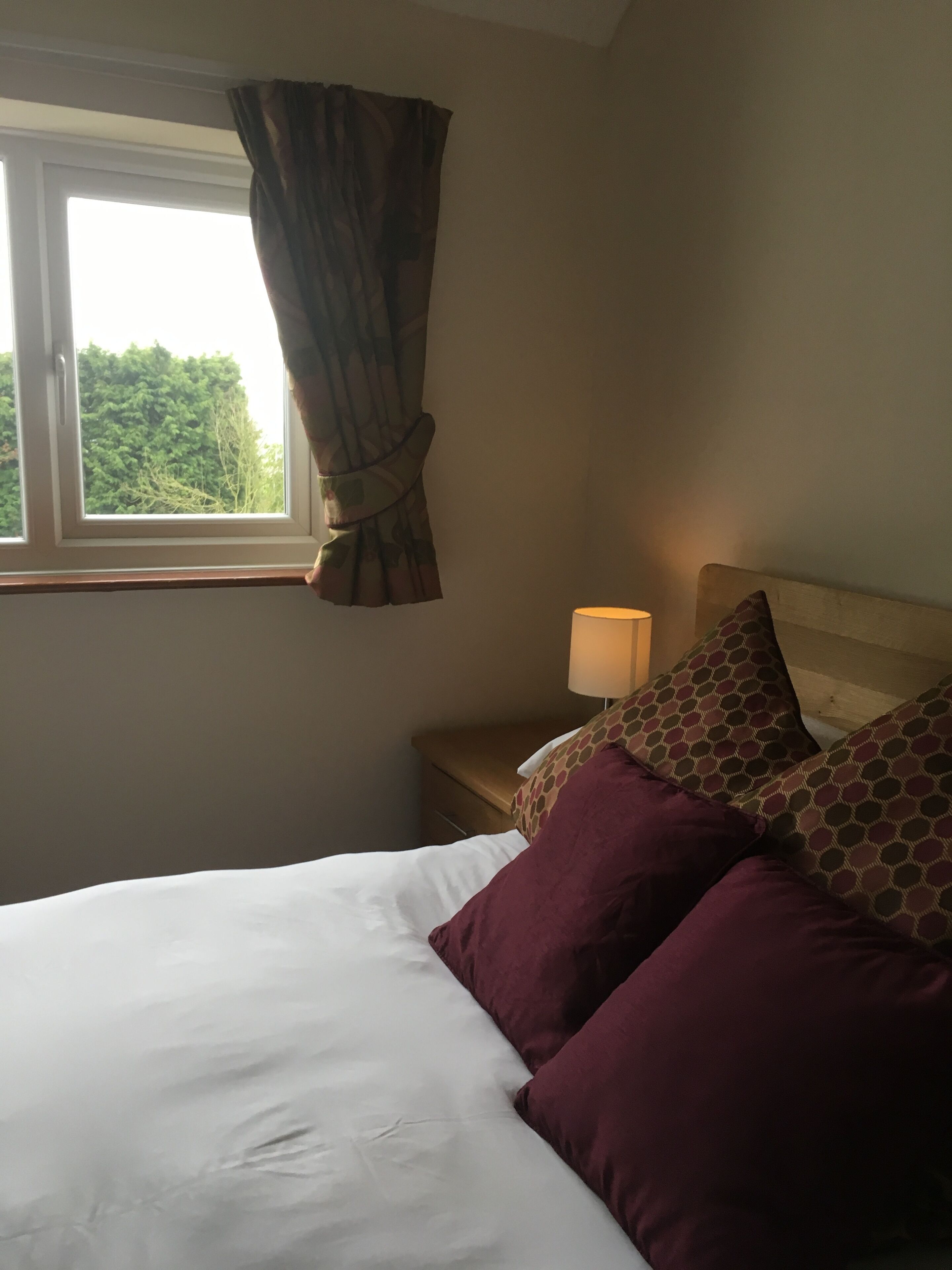 A neatly made bed with white linens and two patterned pillows, a bedside lamp, and a window with curtains.
