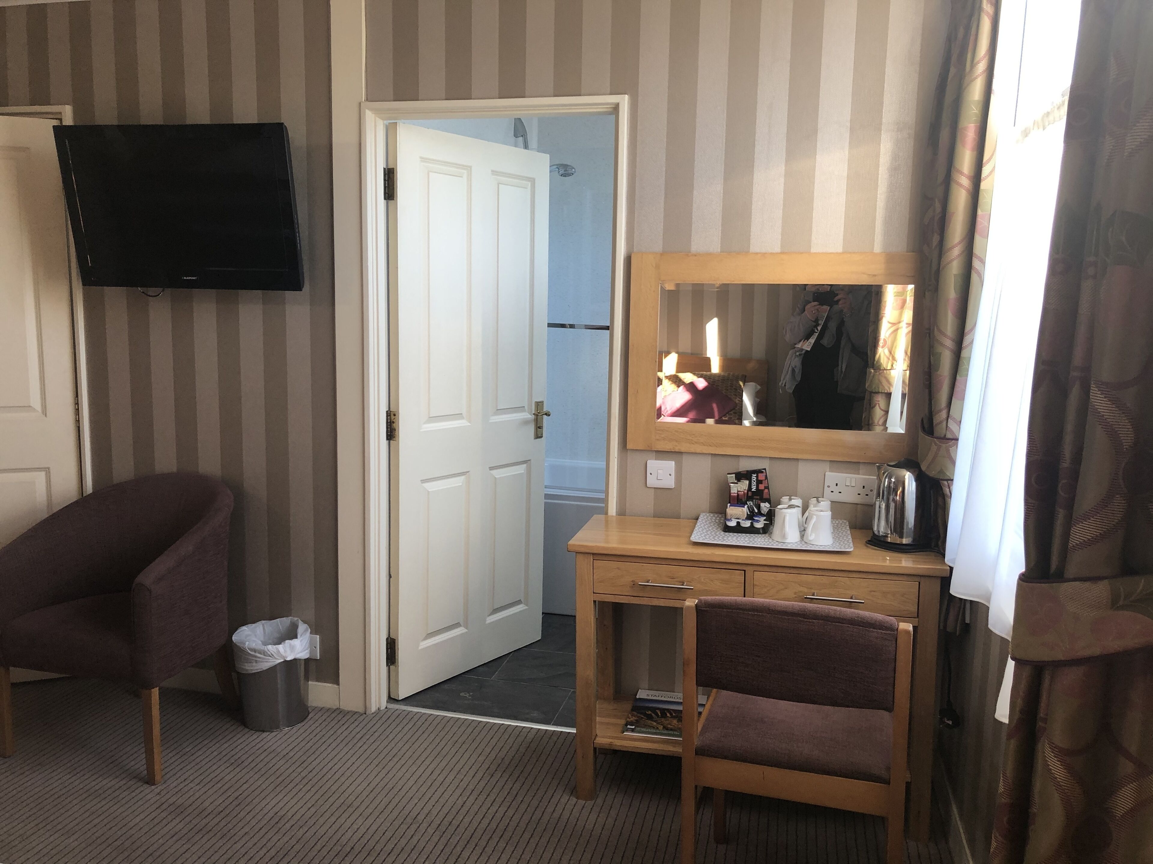 A hotel room with a TV mounted on the wall, a desk with a mirror, a chair, a trash bin, and a door leading to another room.