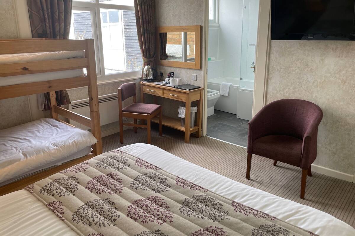 A bedroom with a bunk bed, a desk, a chair, a bed with a floral duvet, a television, and a bathroom visible through an open door.