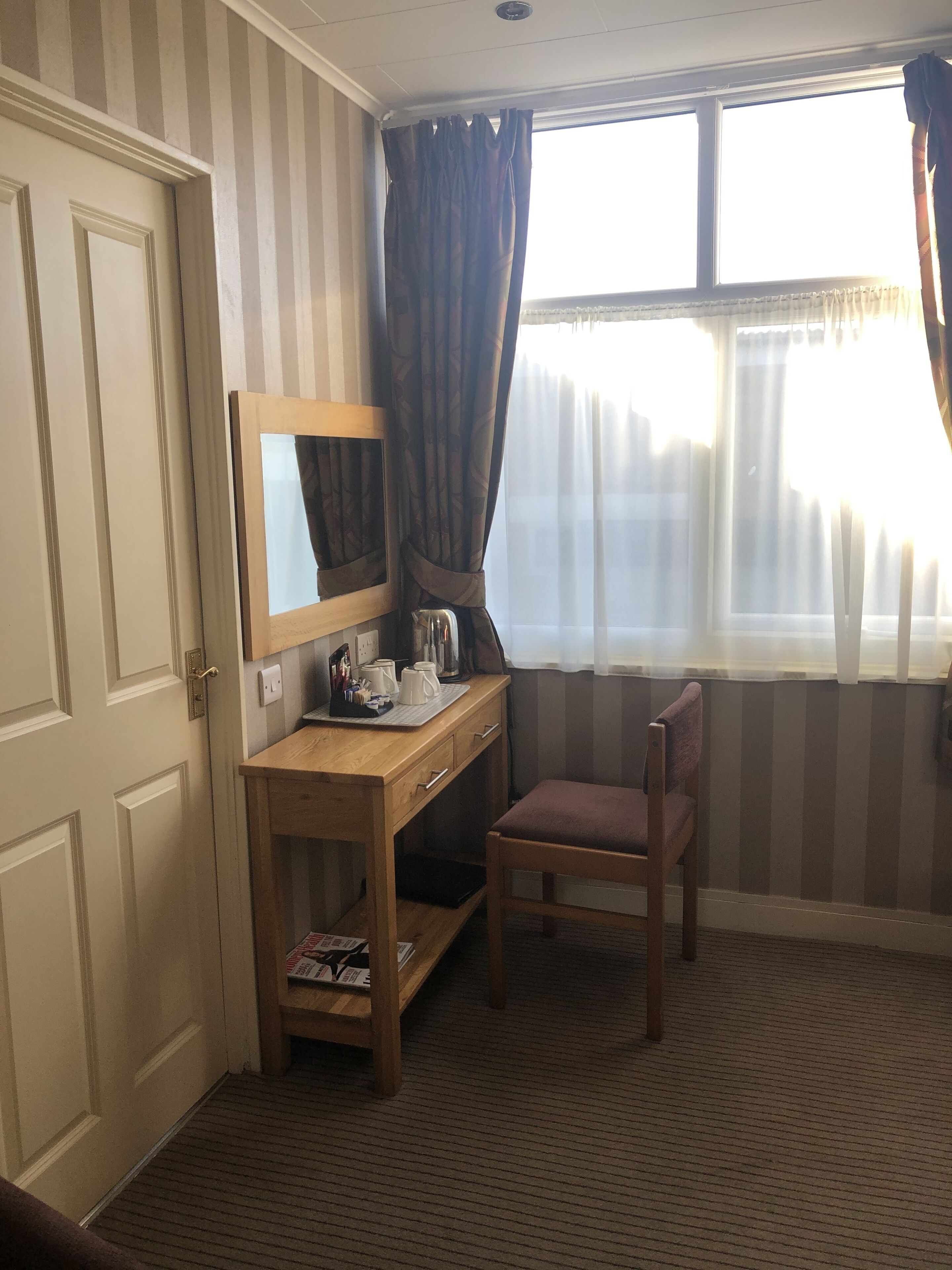 A hotel room with a wooden desk, a chair, a mirror, and a window with curtains.