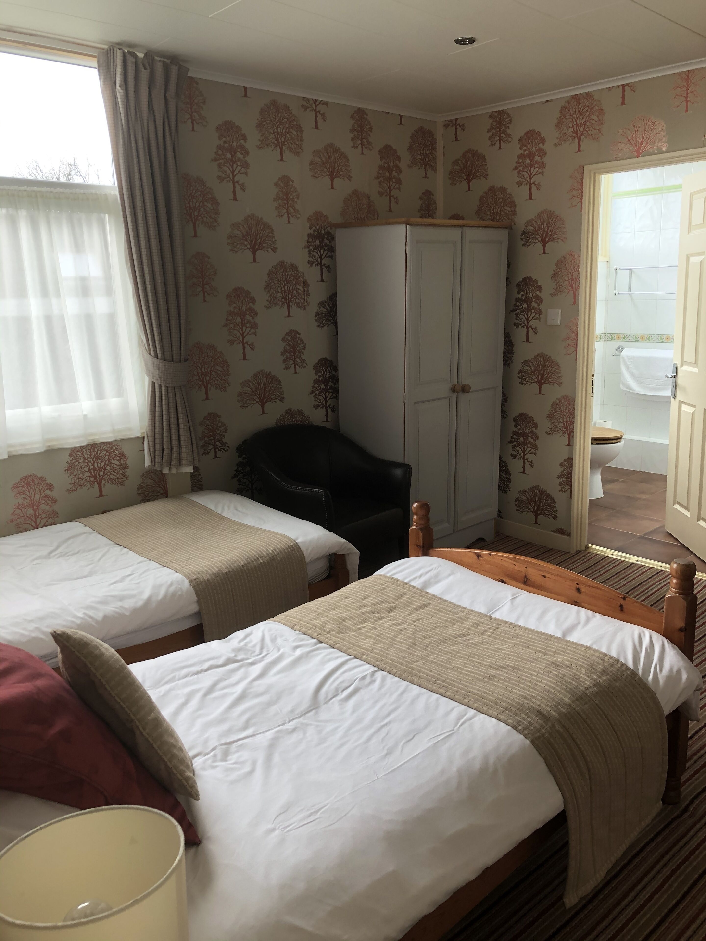 Una habitación de hotel con dos camas individuales, un armario, una silla y un baño visible a través de una puerta abierta.