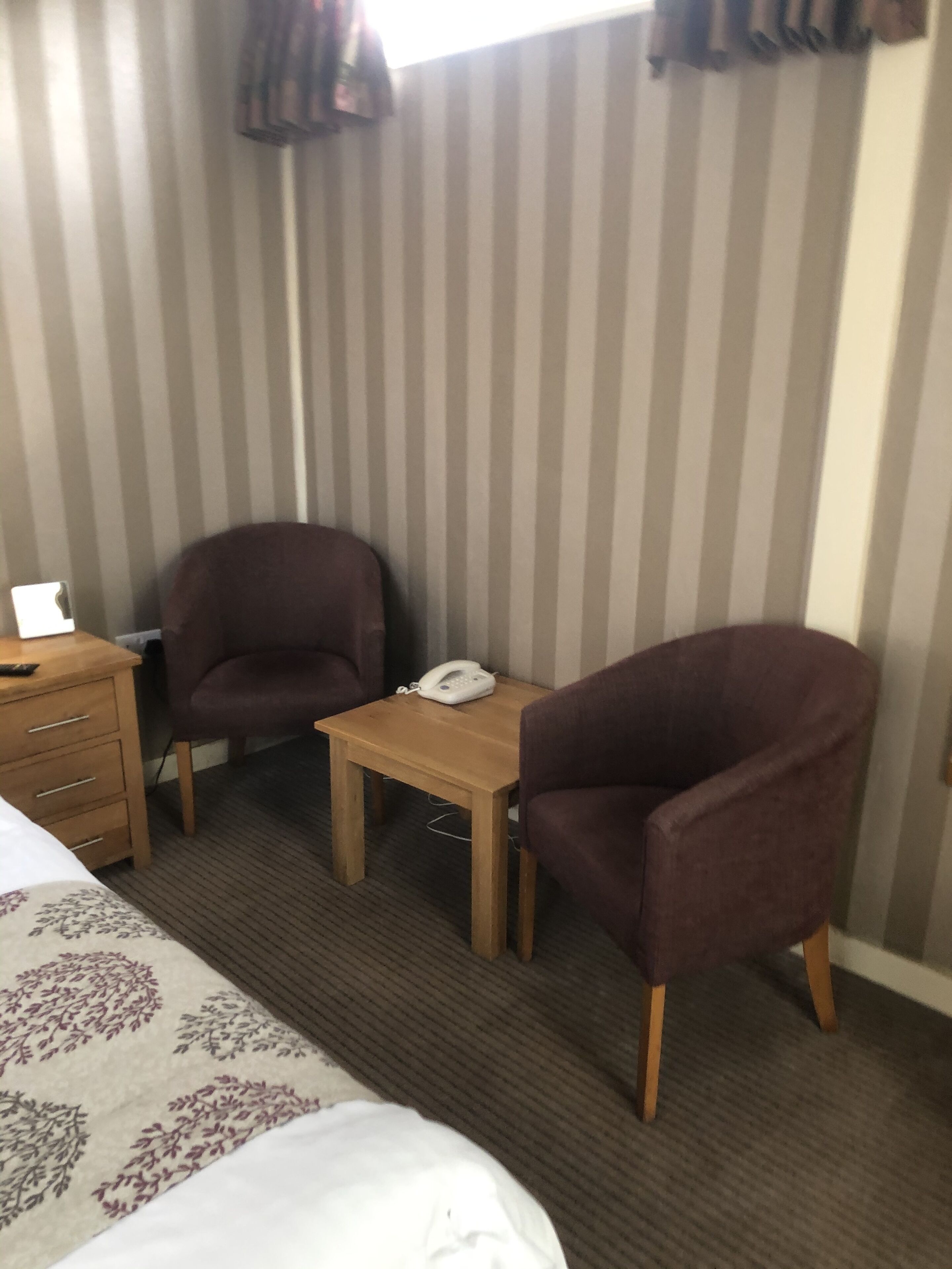 Une chambre d’hôtel avec un lit, deux fauteuils, une table en bois avec un téléphone et un rideau à rayures.