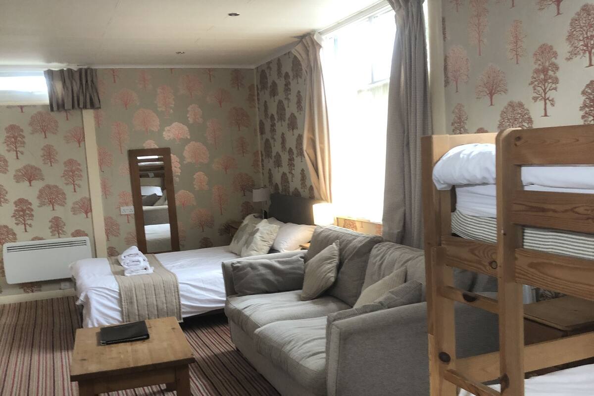 A hotel room with a sofa, two single beds, a wooden bunk bed, a small table, and a mirror.
