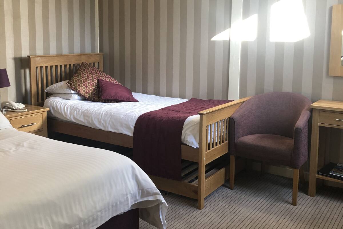 A hotel room with two beds, a wooden headboard, a chair, a nightstand, and a bedside table.