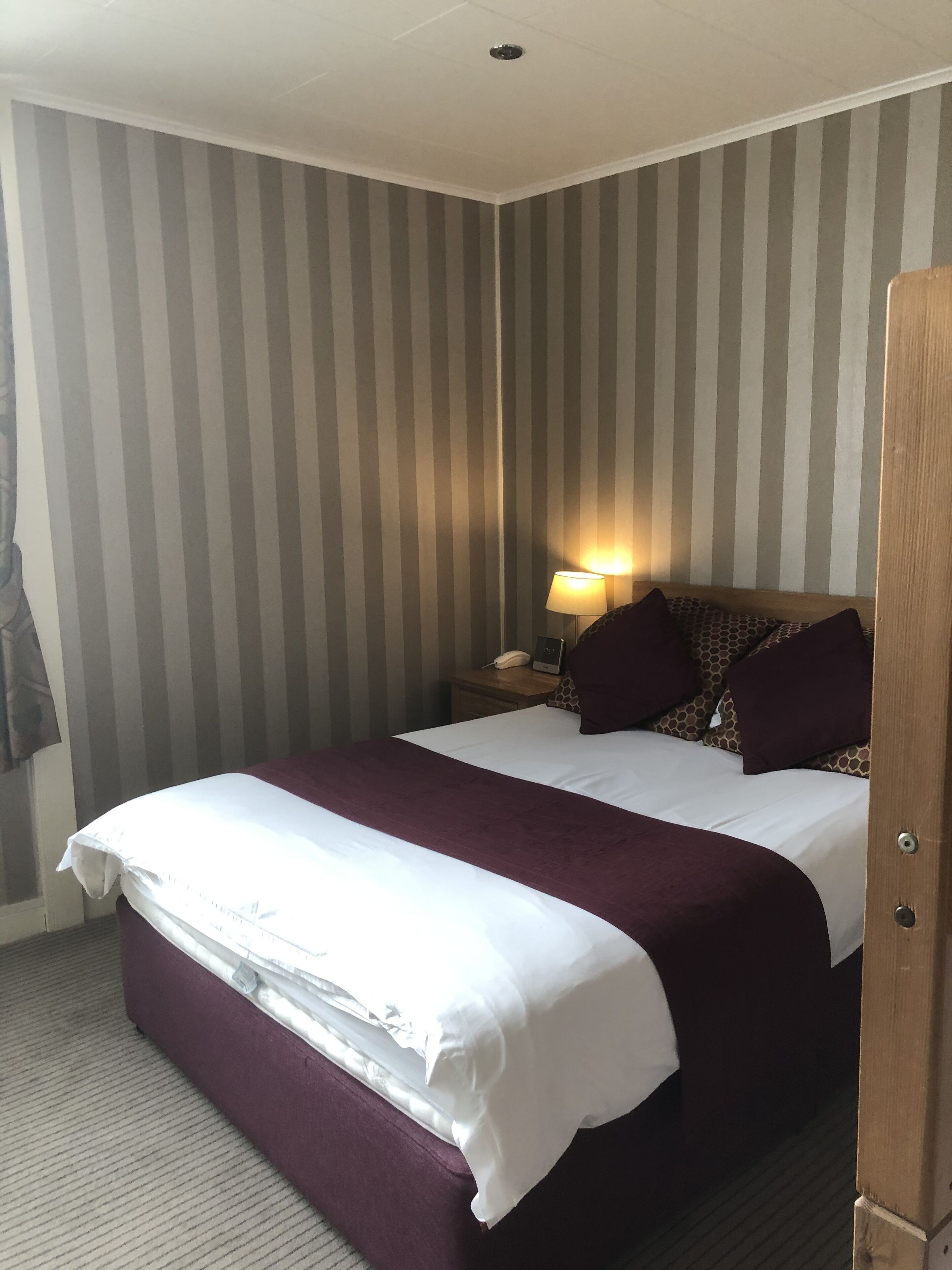 A hotel room with a large bed, striped wallpaper, a nightstand with a lamp, and a patterned headboard.