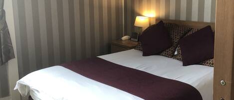 A hotel room with a large bed, striped wallpaper, a nightstand with a lamp, and a patterned headboard.