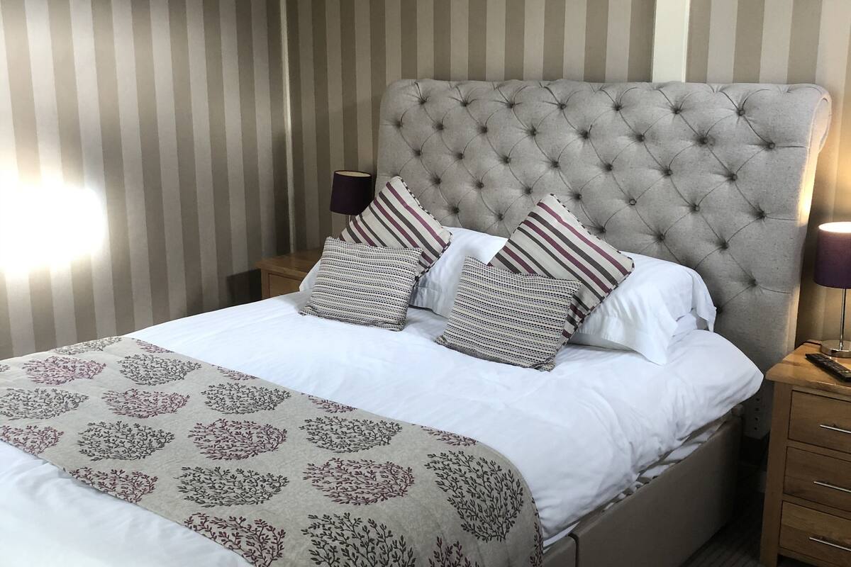 A neatly made bed with a patterned bedspread and pillows, a tufted headboard, and a striped wall in the background.