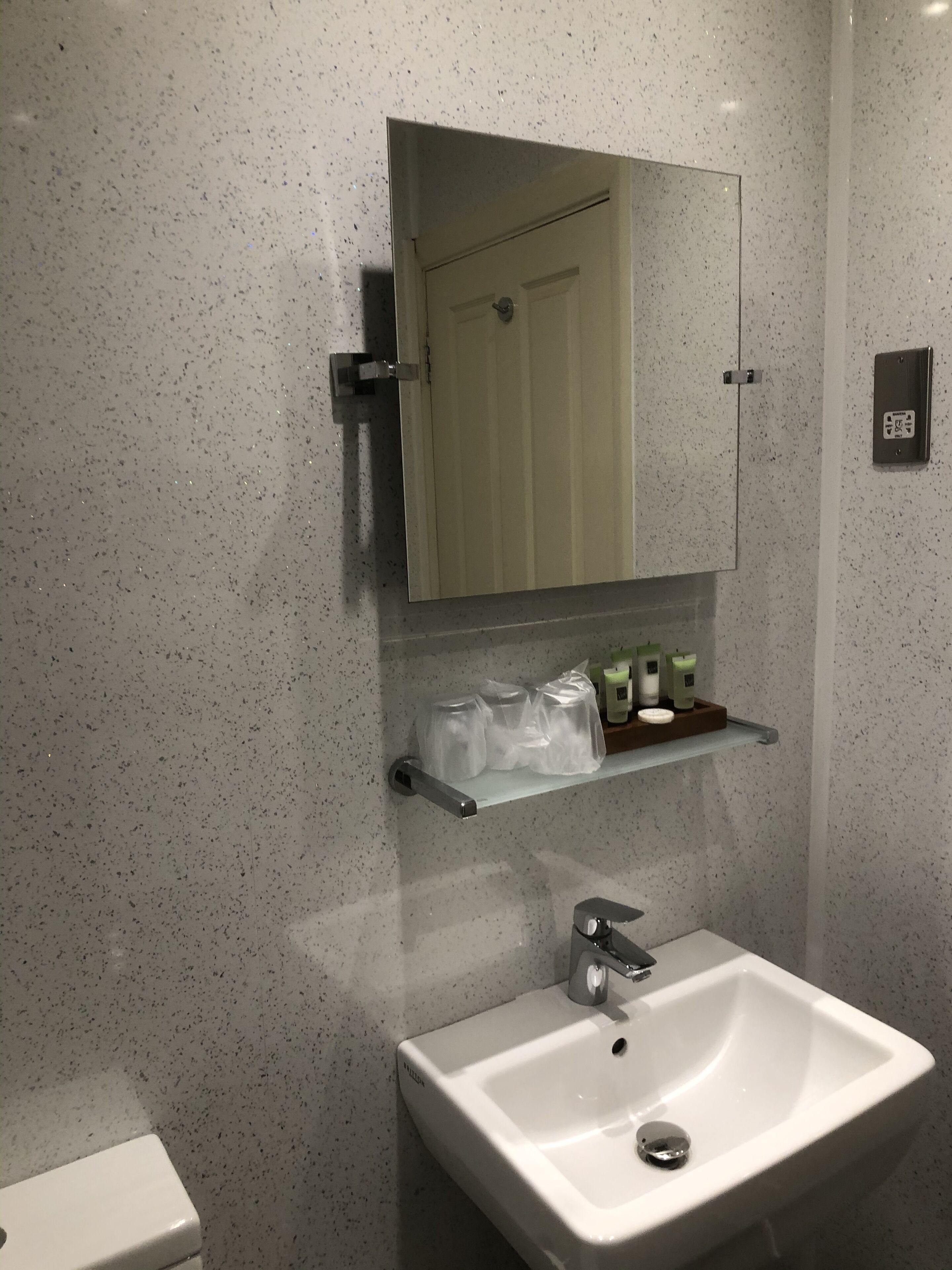 Un baño con un lavamanos blanco, un espejo encima y un estante con artículos de aseo.