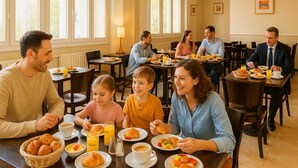 A family dining in a bright, sunlit room with large windows, multiple tables, and a variety of breakfast items. - Hôtel du Parc Montsouris (Paris)