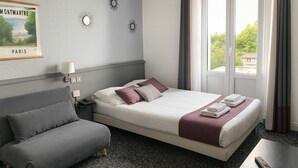 A hotel room with a bed, a sofa, a nightstand, a lamp, a remote control, and a window with curtains. - Hôtel du Parc Montsouris (Paris)