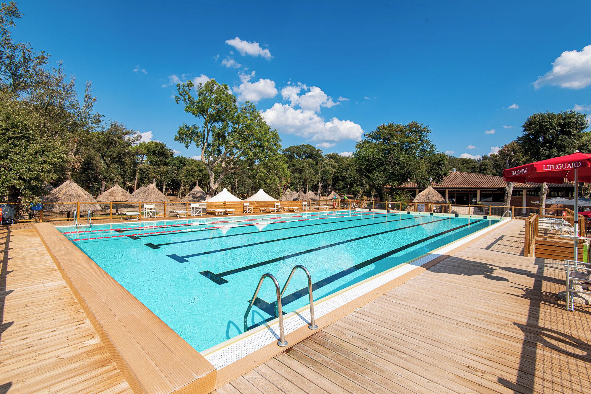 Una piscina con varias pistas de natación, terraza de madera y construcciones con techo de paja.