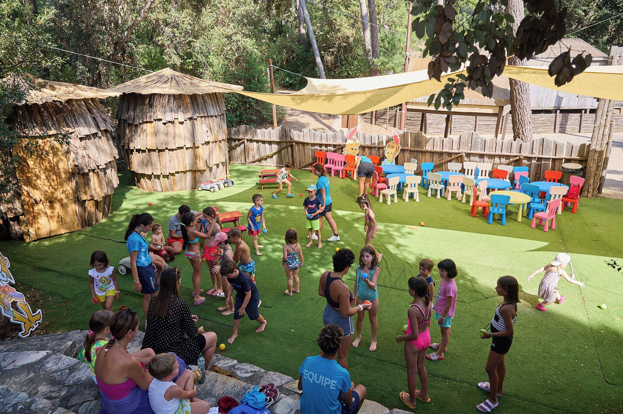 A group of people playing on a grassy area with a pool, surrounded by wooden structures and colorful plastic chairs.