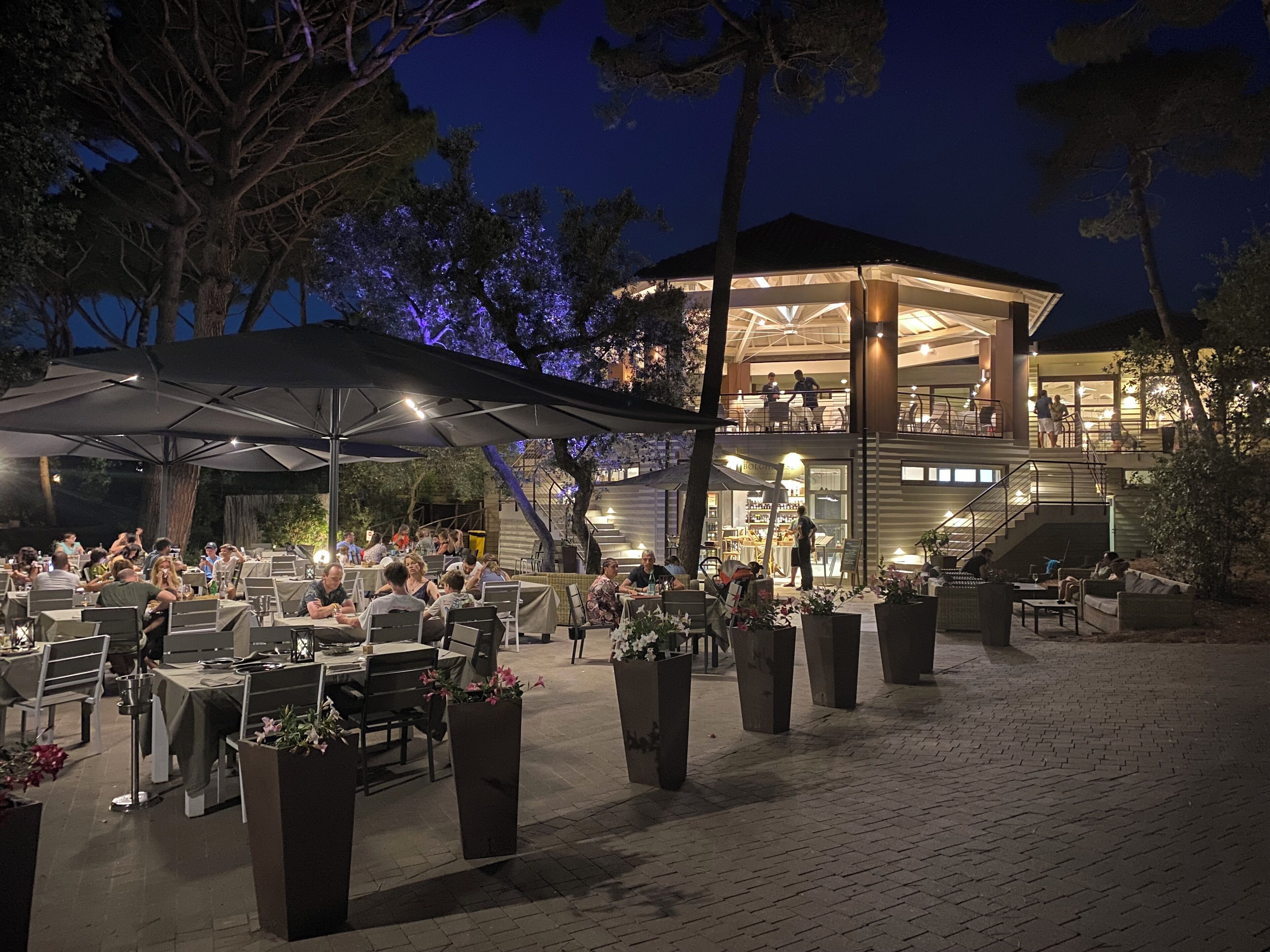 An outdoor dining area with tables and chairs, a covered seating area, and a building with a balcony in the background.