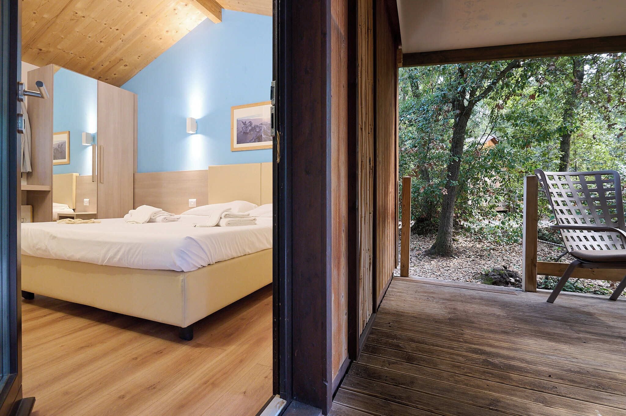 A bedroom with a wooden ceiling, a bed, a nightstand, and a chair by a window overlooking a forest.