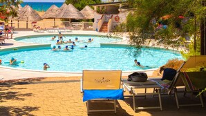 En swimmingpool där folk badar och leker, omgiven av solstolar och parasoller med halmtak.